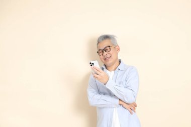 Doğu Asyalı bir adamın portresi gri saçlı ve gözlüklü, şık günlük kıyafetler giymiş, kahverengi arka planda duruyor..