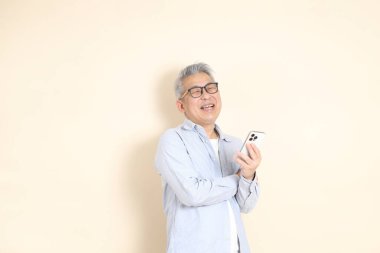 Doğu Asyalı bir adamın portresi gri saçlı ve gözlüklü, şık günlük kıyafetler giymiş, kahverengi arka planda duruyor..