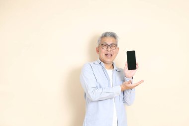 Doğu Asyalı bir adamın portresi gri saçlı ve gözlüklü, şık günlük kıyafetler giymiş, kahverengi arka planda duruyor..