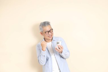 Doğu Asyalı bir adamın portresi gri saçlı ve gözlüklü, şık günlük kıyafetler giymiş, kahverengi arka planda duruyor..