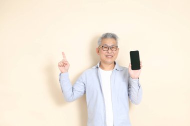 Doğu Asyalı bir adamın portresi gri saçlı ve gözlüklü, şık günlük kıyafetler giymiş, kahverengi arka planda duruyor..