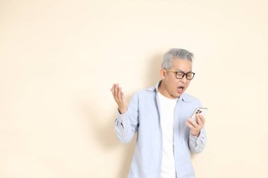 Doğu Asyalı bir adamın portresi gri saçlı ve gözlüklü, şık günlük kıyafetler giymiş, kahverengi arka planda duruyor..