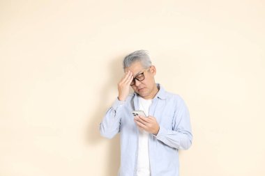 Doğu Asyalı bir adamın portresi gri saçlı ve gözlüklü, şık günlük kıyafetler giymiş, kahverengi arka planda duruyor..