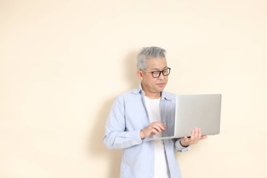 Doğu Asyalı bir adamın portresi gri saçlı ve gözlüklü, şık günlük kıyafetler giymiş, kahverengi arka planda duruyor..