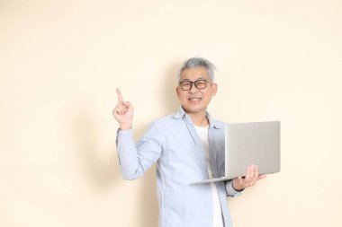 Doğu Asyalı bir adamın portresi gri saçlı ve gözlüklü, şık günlük kıyafetler giymiş, kahverengi arka planda duruyor..