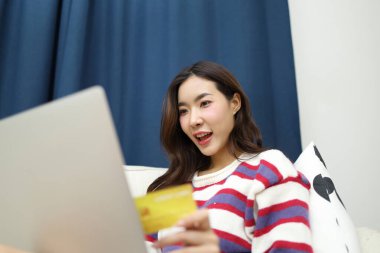 Doğu Asyalı güzel kadın, oturma odasında dizüstü bilgisayarında finansal işler yapmak için bir kredi kartı kullanıyor..