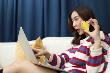 Doğu Asyalı güzel kadın, oturma odasında dizüstü bilgisayarında finansal işler yapmak için bir kredi kartı kullanıyor..