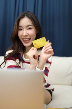 Doğu Asyalı güzel kadın, oturma odasında dizüstü bilgisayarında finansal işler yapmak için bir kredi kartı kullanıyor..