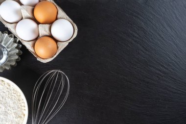 Pişirme malzemeleri kopyalama alanı ile arka plan. Siyah tahta yüzeyde un, yumurta, süt, fırın malzemesi. Üst manzara, düz yatış. Model menüsü, afiş, site başlığı, pişirme kavramı