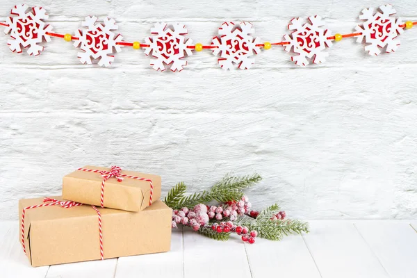 雪の結晶のガーランドと白いレンガの壁の背景に赤い弓とクラフト紙のクリスマスツリーの枝とギフトボックス コピースペースと新年の背景 グリーティングカード — ストック写真