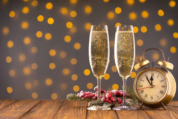 Two wine glasses with bubbly champagne, fir branch with decor and bronze clock on background of blurry sparkling lights. Happy New Year holiday greeting card, banner, header with copy space