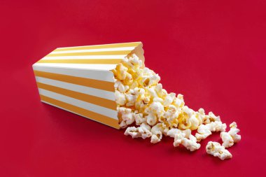 Kırmızı arka planda izole edilmiş sarı çizgili bir karton kovadan düşen lezzetli peynirli patlamış mısır. Patlamış mısır taneleri saçılıyor. Sinema, sinema ve eğlence konsepti.
