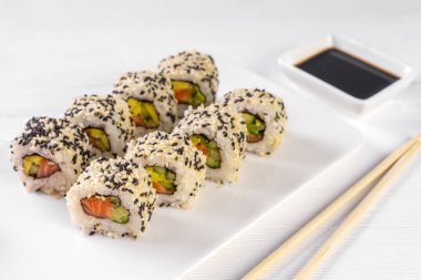 Sushi roll California with salmon, sesame seeds, avocado, cream cheese on white background. Sushi menu, rolls served on white dish and soy sauce. Traditional Japanese cuisine food.