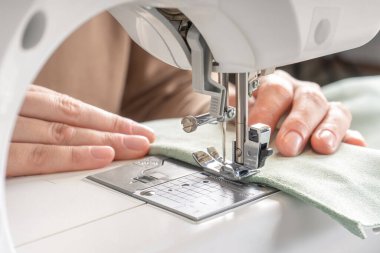 Atölyedeki iş yerindeki modern dikiş makinesine beyaz kumaş diken kadın elleri. Kadın eli, dikiş makinesine kumaş parçaları dikiyor. El yapımı, hobi, küçük işletme konsepti