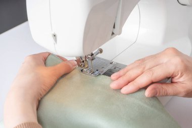 Atölyedeki iş yerindeki modern dikiş makinesine beyaz kumaş diken kadın elleri. Kadın elleri dikiş makinesinin üzerine kumaş parçaları dikiyor. El yapımı, hobi, küçük işletme konsepti