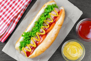 Sarı hardallı, ketçaplı, domatesli ve siyah arkaplanlı taze salata yapraklı ev yapımı Hot Dog. Fast food, sokak yemeği, Amerikan mutfağı.