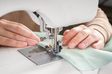 Atölyedeki iş yerinde modern dikiş makinesine yeşil kumaş diken kadın elleri. Kadın elleri dikiş makinesinin üzerine kumaş parçaları dikiyor. El yapımı, hobi, küçük işletme konsepti