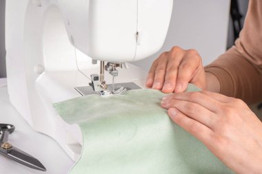 Atölyedeki modern dikiş makinesine yeşil kumaş diken terzi elleri. Kadın elleri dikiş makinesinin üzerine kumaş parçaları dikiyor. El yapımı, hobi, onarım, küçük işletme konsepti