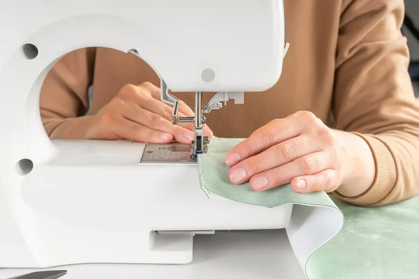 アトリエの職場で現代ミシンでグリーンファブリックをステッチするテーラーハンド 女性の手は縫製機のクローズアップに布の部分を縫います ハンドメイド 中小企業コンセプト — ストック写真