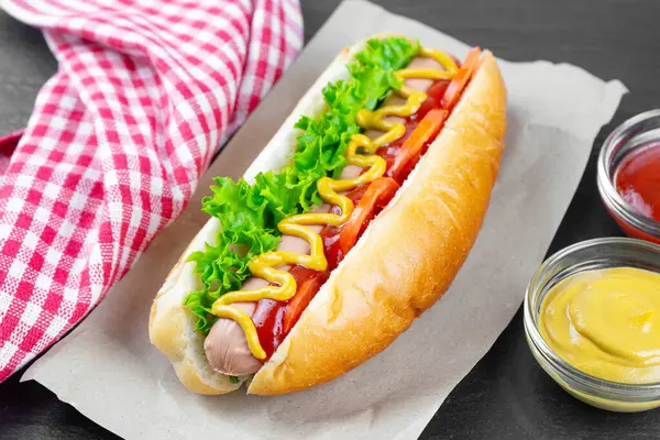 Homemade Hot Dog with yellow mustard, ketchup, tomato and fresh salad leaves on black slate background. Fast food, street food, american cuisine