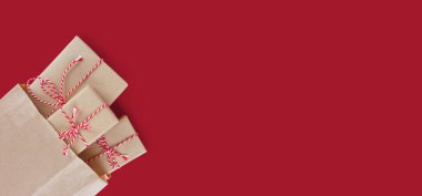 Noel hediyesi kutuları el işi kağıdına sarılıp kırmızı arka planda kırmızı ve beyaz pastacı ipiyle kahverengi geri dönüşümlü kağıt torbaya doldurulmuş. Düz yatış, üst görünüm, afiş, başlık.