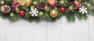 Altın ve yeşil toplarla süslenmiş Noel ağacı dalları, beyaz ahşap arka planda kırmızı bereler ve kar taneleri. Üst görünüm, düz görünüm kopyalama alanı, afiş, başlık, Yeni Yıl arkaplanı