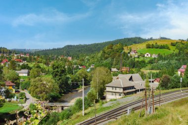 Vadideki güzel köy manzarası ve yaz günü dağların arasında demiryolu. Bulutlu mavi gökyüzü, orman tepeleri, çam ve çam ağaçları, kırsal huzur manzarası. Karpatların Doğası, Ukrayna.