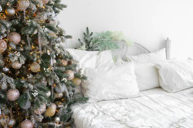 Tatiller için süslenmiş rahat yatak odası köşesi altın süslerle süslenmiş Noel ağacı, beyaz yatak ve yumuşak yastıklı çelenkler, rahat bir tatil ortamı yaratıyor..
