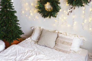 Yatak odasında Noel ağacı ve çelenk, kış aylarında tatil dekorasyonu ve sıcak aydınlatma. Beyaz çarşaflı yatak ve yumuşak dekoratif yastıklar. Kış tatilleri için sıcak, şenlikli bir atmosfer..