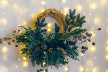 Çam dallarından yapılmış güzel bir Noel çelengi ve beyaz duvardaki dekoratif elementlerden yapılmış yumuşak, sıcak peri ışıklarıyla aydınlatılmış. Kış tatili atmosferi için ev dekorasyonu.
