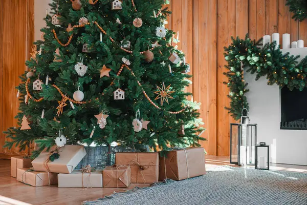 Güzel süslenmiş Noel ağacı şöminenin yanında duruyor. Odanın arka planında, sarılı hediye kutularıyla süslenmiş çelenklerle süslenmiş. Tatil atmosferi için şenlikli ev dekorasyonu.