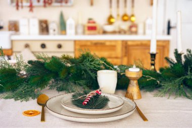 Saf mutfak arka planında yeşil ve basit dekorlu zarif bir Noel sofrası. Tabaklar, mumlar ve beyaz masa örtüsü üzerinde altın kültürü olan güzel düzenlenmiş tatil masası.