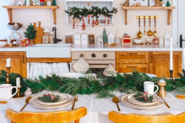 Saf mutfak arka planında yeşil ve basit dekorlu zarif bir Noel sofrası. Tabaklar, mumlar ve beyaz masa örtüsü üzerinde altın kültürü olan güzel düzenlenmiş tatil masası.