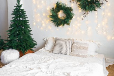 Yatak odasında Noel ağacı ve çelenk, kış aylarında tatil dekorasyonu ve sıcak aydınlatma. Beyaz çarşaflı yatak ve yumuşak dekoratif yastıklar. Kış tatilleri için sıcak, şenlikli bir atmosfer..