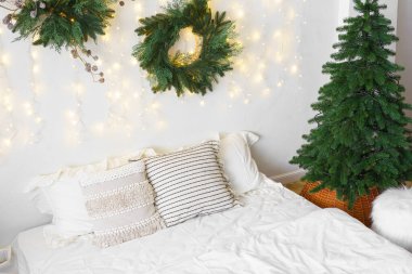 Yatak odasında Noel ağacı ve çelenk, kış aylarında tatil dekorasyonu ve sıcak aydınlatma. Beyaz çarşaflı yatak ve yumuşak dekoratif yastıklar. Kış tatilleri için sıcak, şenlikli bir atmosfer..