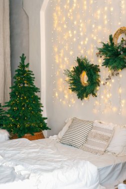 Yatak odasında Noel ağacı ve çelenk, kış aylarında tatil dekorasyonu ve sıcak aydınlatma. Beyaz çarşaflı yatak ve yumuşak dekoratif yastıklar. Kış tatilleri için sıcak, şenlikli bir atmosfer..