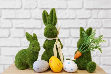 Trhee dekoratif yeşil yosunlu tavşan figürleri. Beyaz tuğlalı arka planda ahşap masa üzerinde renkli Paskalya yumurtaları. Paskalya bayramı atmosferi, duvar kağıdı, fon.