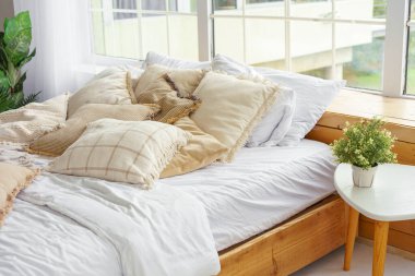 Yatak odasındaki yumuşak yastıklı rahat yatak penceresinde doğal ışık var. Tarafsız tonlarda yumuşak yastıklarla süslenmiş rahat bir yatak. Boho tarzında tasarım elementi