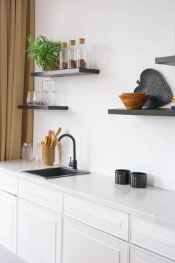 Tezgahı beyaz mutfak ve beyaz duvar arkasında siyah su musluğu ve ahşap rafları var. Mutfak, mutfak ve dekorlu raflar. İç tasarım, minimalist stil
