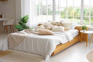 Açık güneşli yatak odasında yeşil bitkiler ve geniş pencereli yumuşak bej yastıklı rahat bir yatak. Boho tarzında oturma odasının iç tasarımı