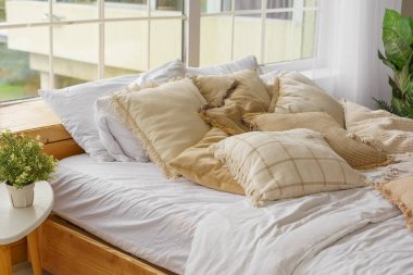 Yatak odasındaki yumuşak yastıklı rahat yatak penceresinde doğal ışık var. Tarafsız tonlarda yumuşak yastıklarla süslenmiş rahat bir yatak. Boho tarzında tasarım elementi