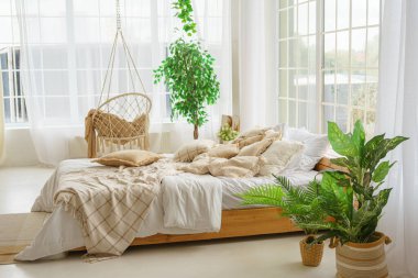 Açık güneşli yatak odasında yeşil bitkiler ve geniş pencereli yumuşak bej yastıklı rahat bir yatak. Boho tarzında oturma odasının iç tasarımı