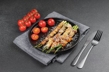 Kızarmış kuşkonmaz, üzerine parmesan peyniri serpiştirilmiş ve siyah arka planda taze vişneli domatesle servis edilmiş domuz pastırması. Gurme yemek, yemek tarifi, mutfak veya restoran menüsü