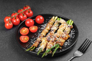 Kızarmış kuşkonmaz, üzerine parmesan peyniri serpiştirilmiş ve siyah arka planda taze vişneli domatesle servis edilmiş domuz pastırması. Gurme yemek, yemek tarifi, mutfak veya restoran menüsü