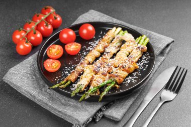 Kızarmış kuşkonmaz, üzerine parmesan peyniri serpiştirilmiş ve siyah arka planda taze vişneli domatesle servis edilmiş domuz pastırması. Gurme yemek, yemek tarifi, mutfak veya restoran menüsü