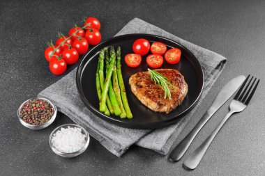 Lezzetli ızgara biftek, yeşil kuşkonmaz ve siyah tabakta, çatal bıçakla servis edilen kiraz domatesleri. Yemek menüsü, diyet ya da mutfak tasarımı için mükemmel..