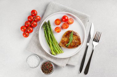 Lezzetli ızgara biftek, yeşil kuşkonmaz ve beyaz tabakta vişneli domates, çatal bıçak ve baharatla servis ediliyor. Yemek menüsü, diyet ya da mutfak tasarımı için mükemmel..