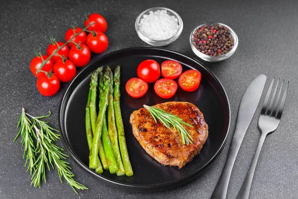 Lezzetli ızgara biftek, yeşil kuşkonmaz ve siyah tabakta, çatal bıçakla servis edilen kiraz domatesleri. Yemek menüsü, diyet ya da mutfak tasarımı için mükemmel..