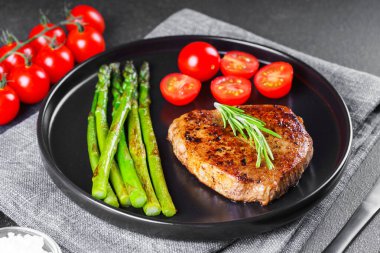 Yaklaş, yeşil kuşkonmaz ve siyah tabak üzerinde vişneli domatesle servis edilen lezzetli ızgara biftek makrosu. Yemek menüsü, diyet ya da mutfak tasarımı için mükemmel..