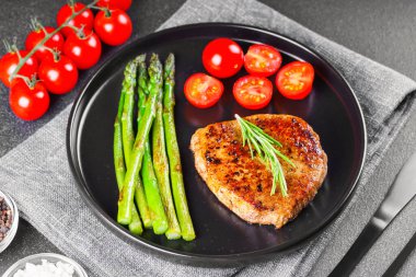 Lezzetli ızgara biftek, yeşil kuşkonmaz ve siyah tabakta, çatal bıçakla servis edilen kiraz domatesleri. Yemek menüsü, diyet ya da mutfak tasarımı için mükemmel..